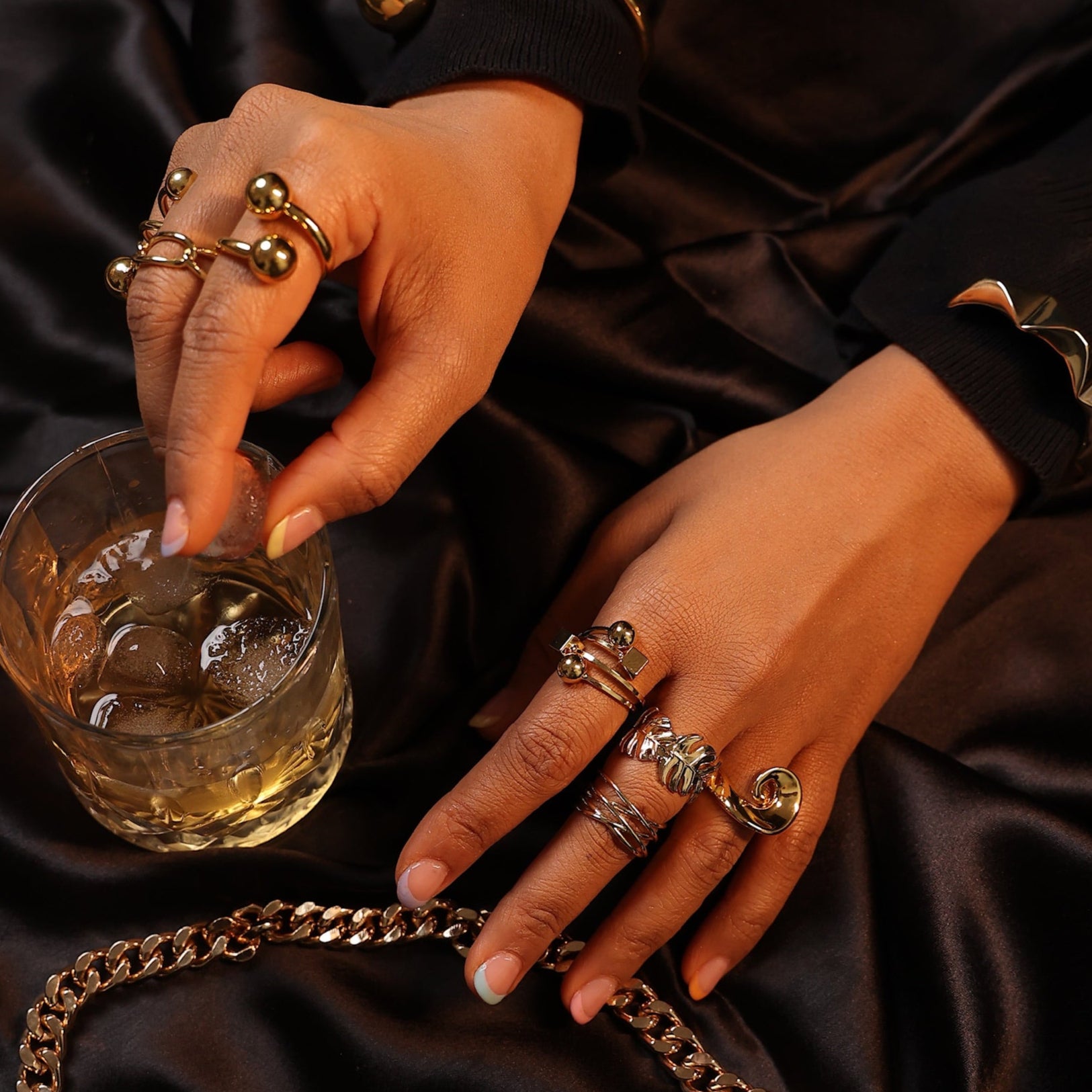 Close-up of hands with gold rings holding a glass of whiskey on a dark fabric background