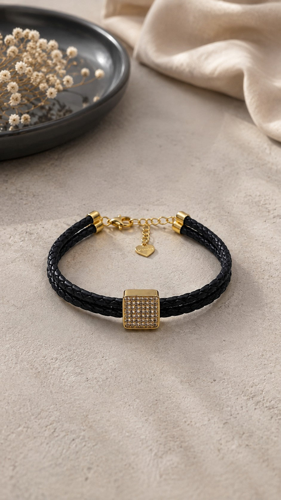 Daga-accessories black braided cord bracelet featuring a gold-toned square crystal charm and heart clasp detail.