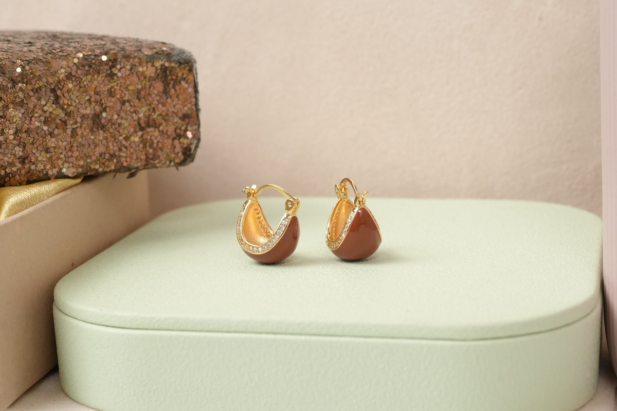 Daga-accessories brown enamel huggie hoop earrings with gold-toned metal and cubic zirconia accents on a green stand.