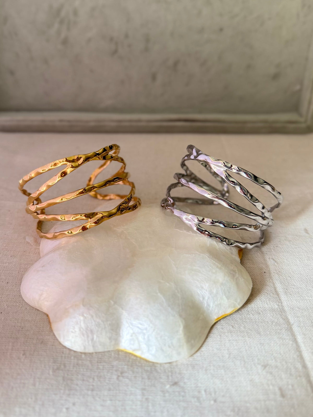Daga-accessories gold and silver criss-cross metal cuffs with a textured hammered finish on a white decorative base.