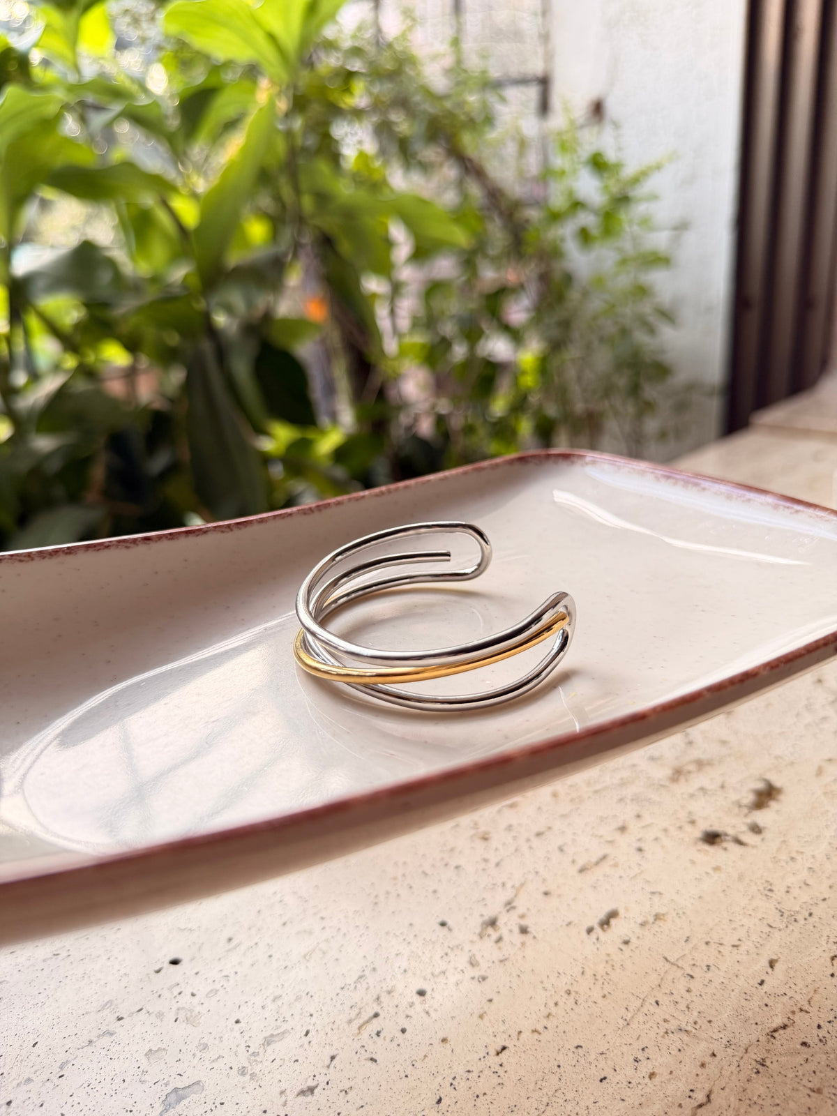 Daga-accessories dual tone arc cuff bracelet featuring a curved silver and gold metallic design on a ceramic tray.