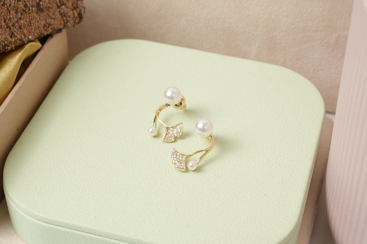 Daga-accessories gold-toned ginkgo leaf stud earrings with faux pearls and micro-crystals on a light green display.
