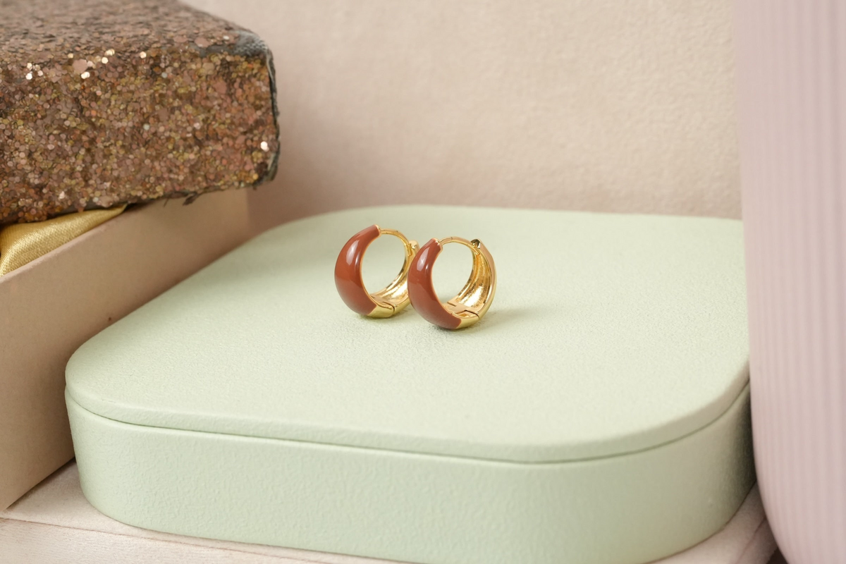 Daga-accessories gold-toned huggie hoop earrings with a glossy brown enamel inlay on a light green display stand.