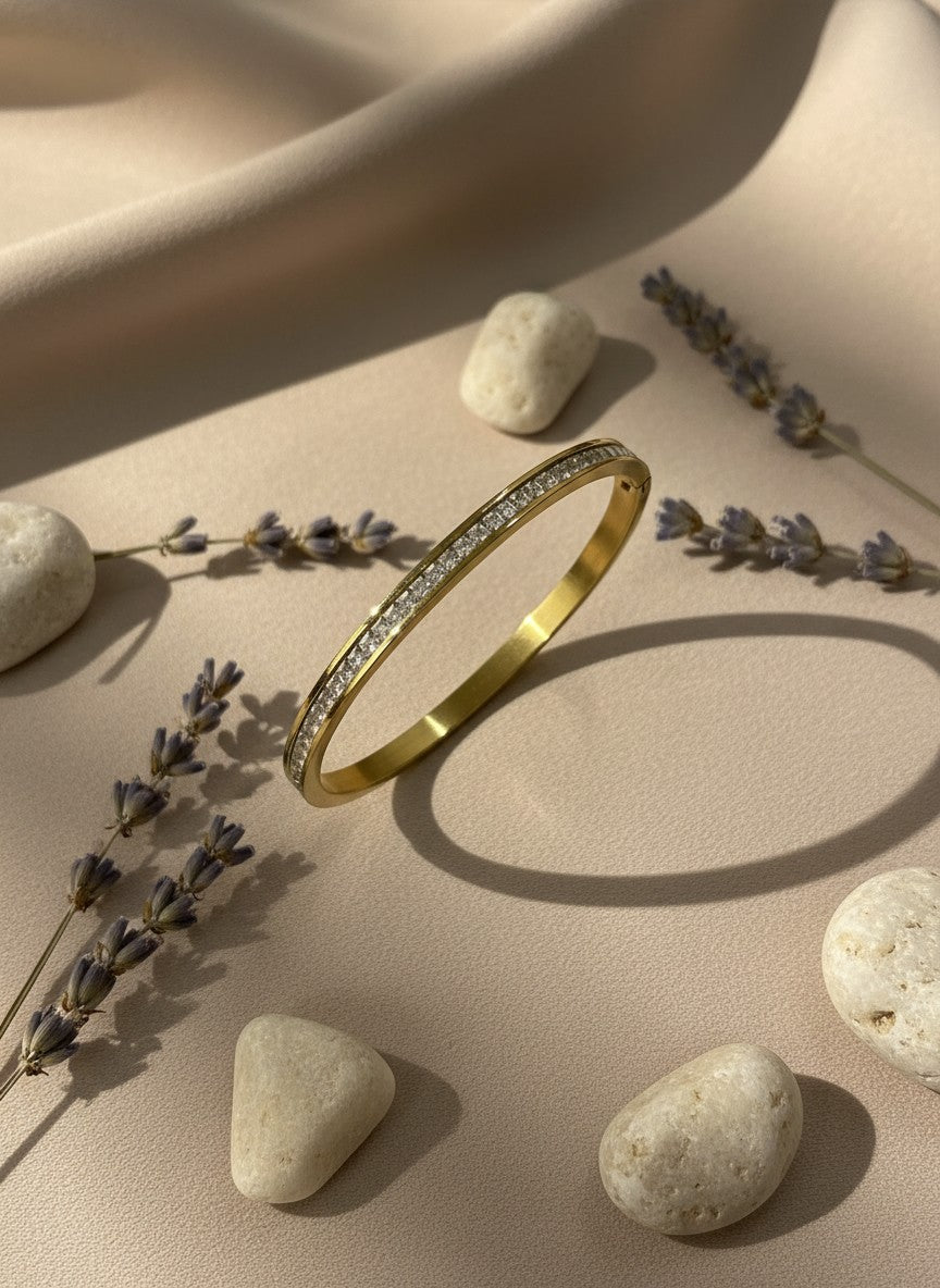 Daga-accessories gold-toned bangle bracelet featuring a row of clear crystals on a soft beige fabric background.