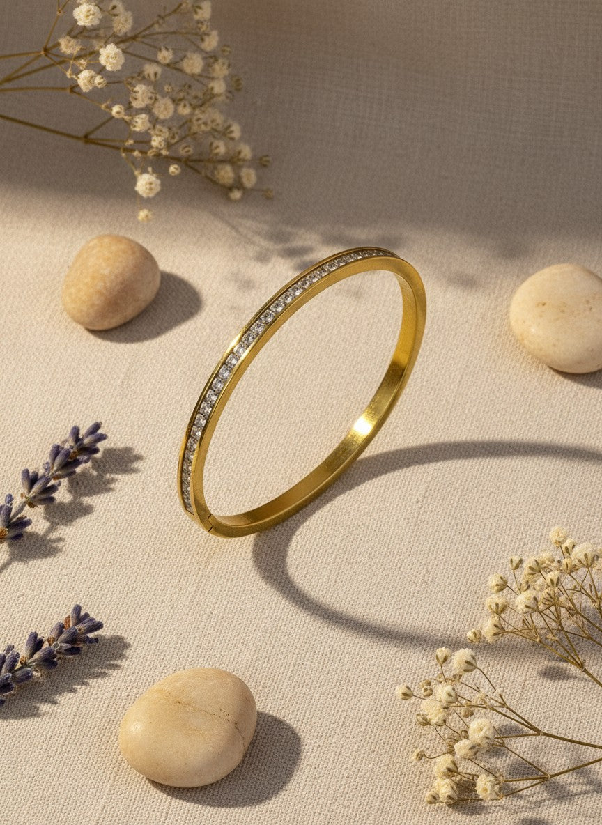Daga-accessories gold-toned metal bangle bracelet featuring a row of clear crystals on a textured beige background.