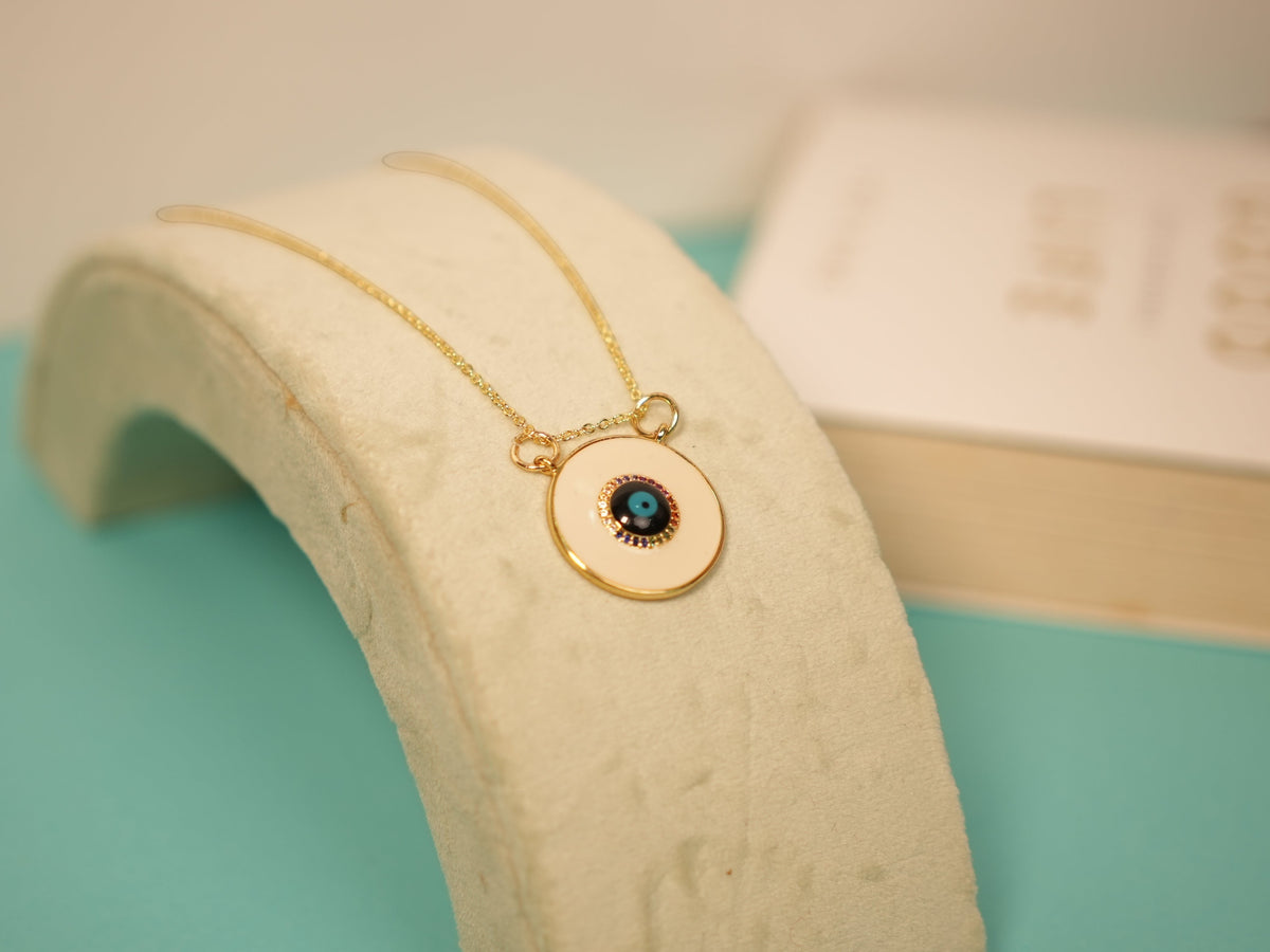 Daga-accessories gold-tone necklace with a cream enamel disc pendant featuring a blue and black evil eye detail.