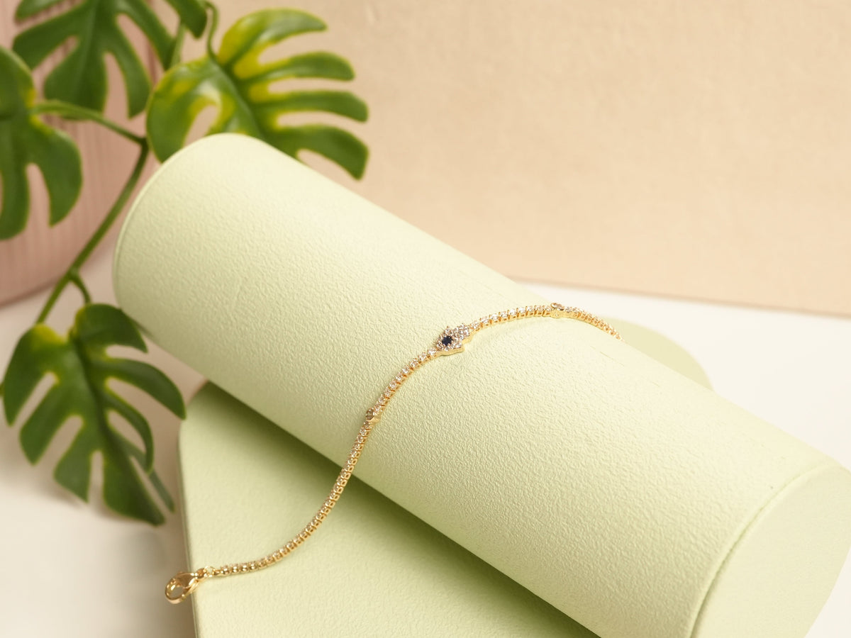 Daga-accessories gold tennis bracelet featuring a Hamsa hand charm with a blue center stone on a light green display