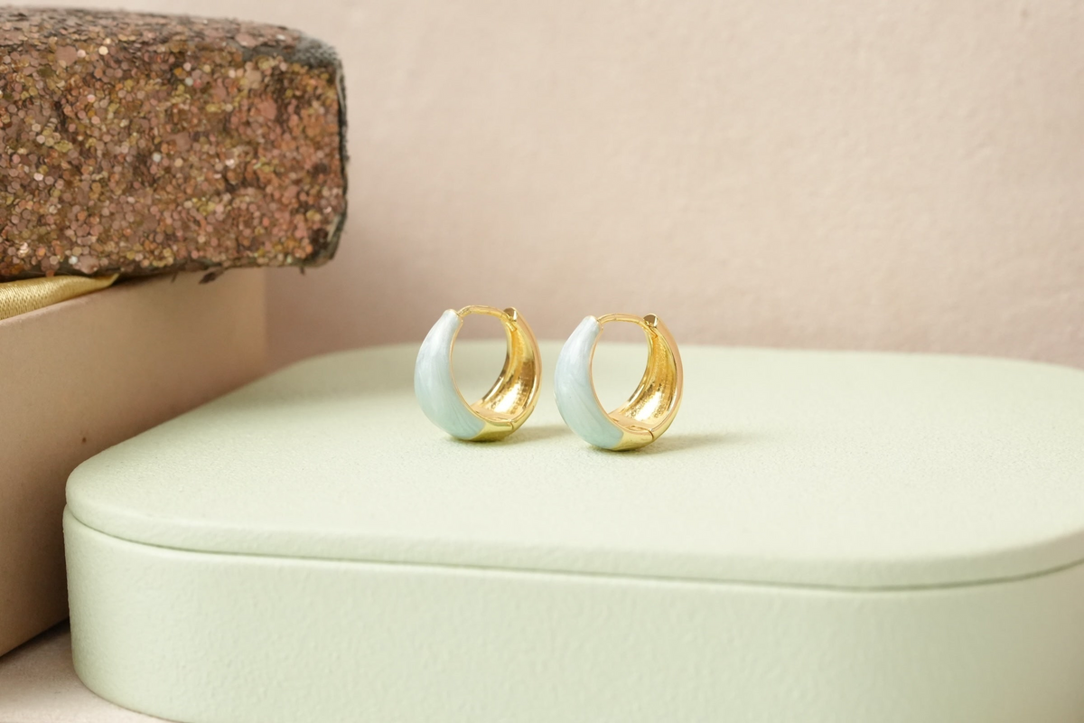 Daga-accessories gold-plated huggie hoop earrings with pastel mint enamel inlay on a soft green display stand.