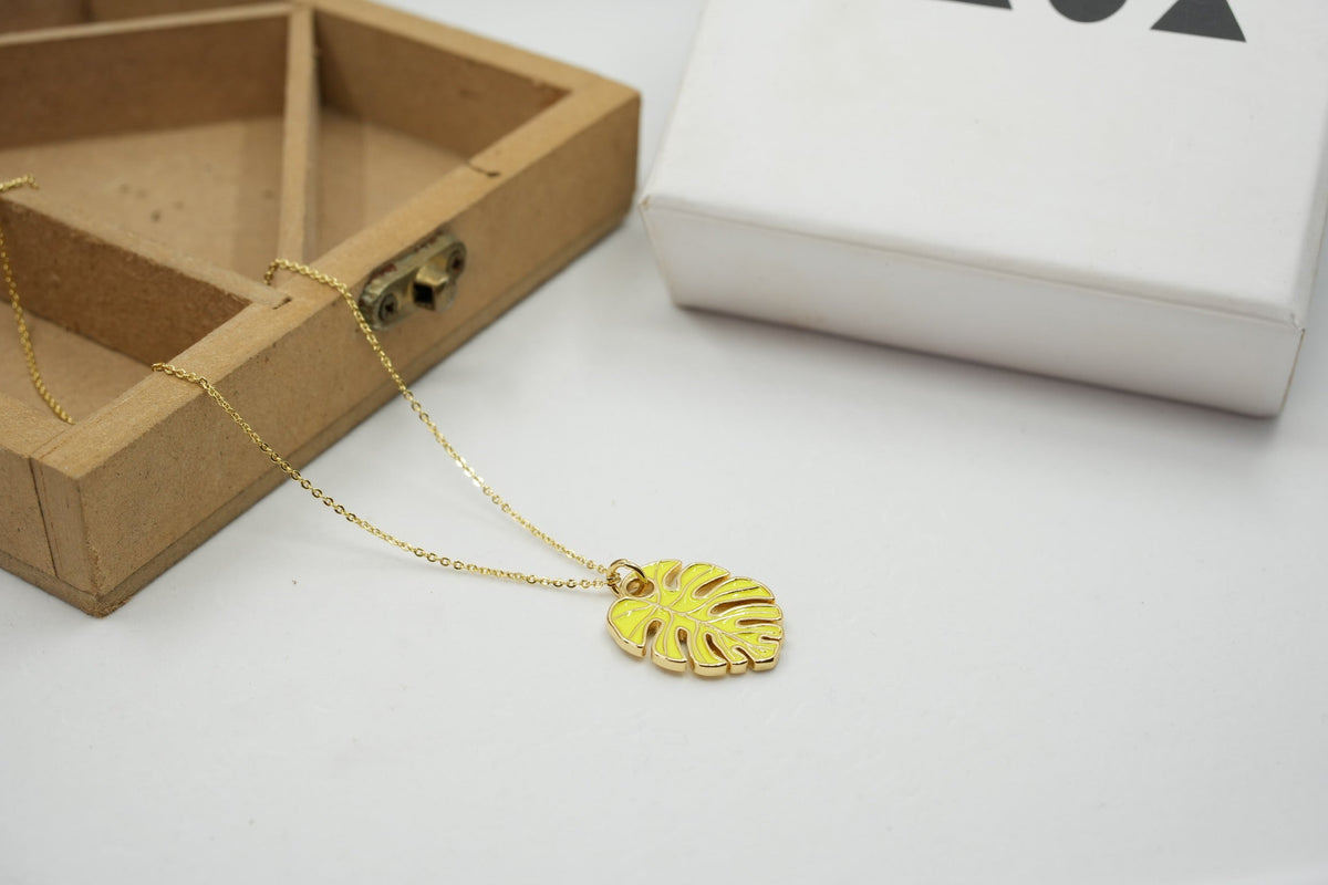 Daga-accessories gold chain necklace featuring a yellow monstera leaf pendant on a white surface with a wooden box