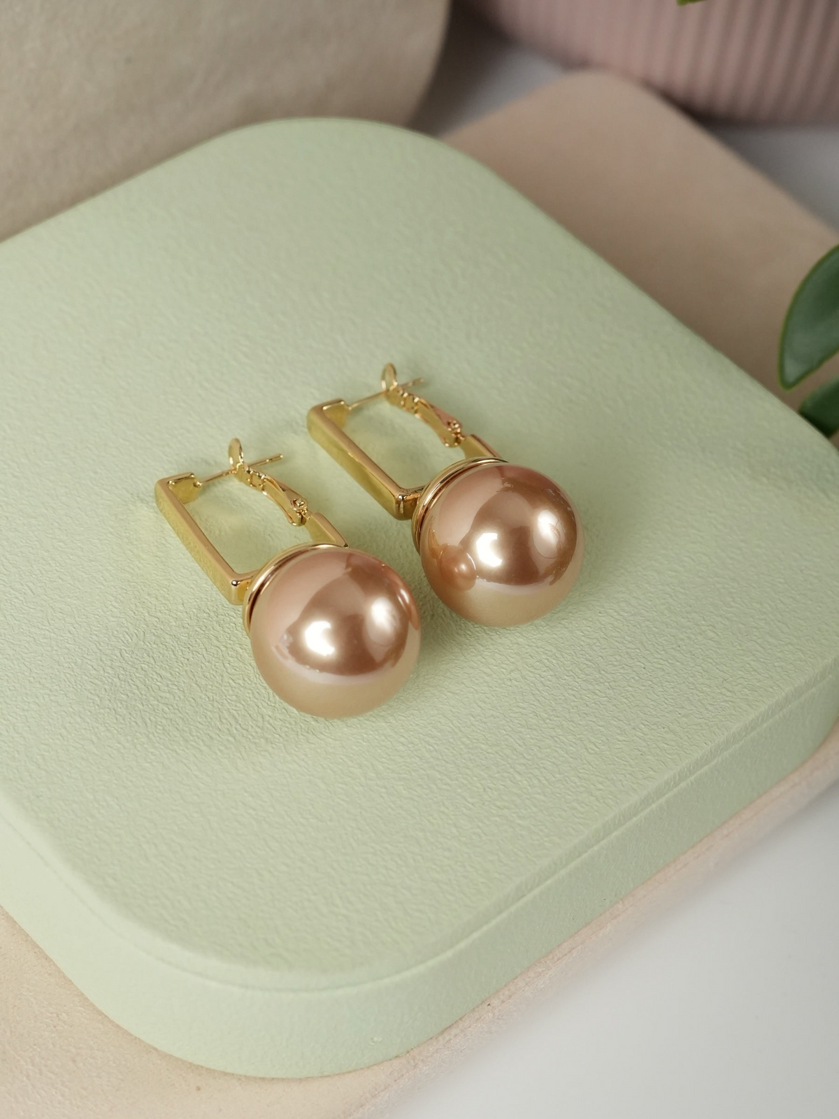 Daga-accessories gold-toned rectangular hoop earrings featuring large lustrous spherical pearls on a green display base.