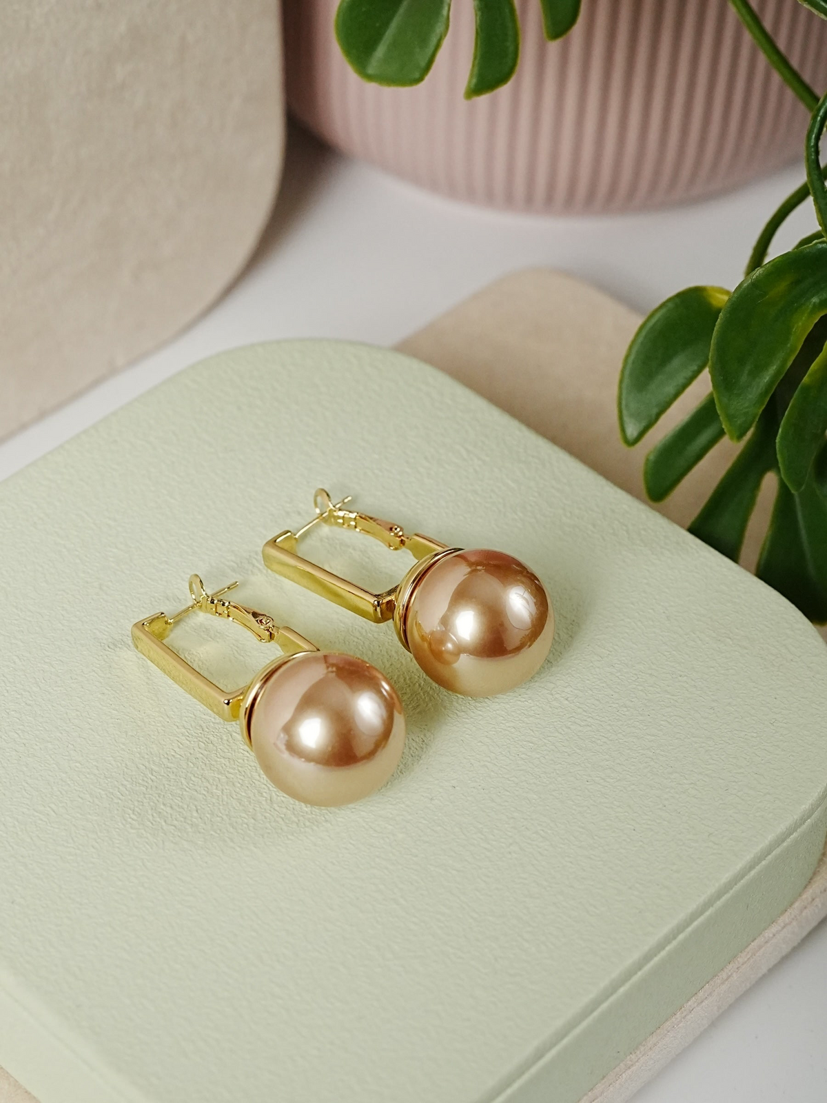 Daga-accessories gold-toned rectangular hoop earrings featuring large lustrous spherical pearls on a green display.