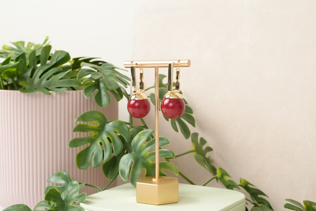 Daga-accessories gold-toned drop earrings with vibrant red spherical charms displayed on a gold stand.