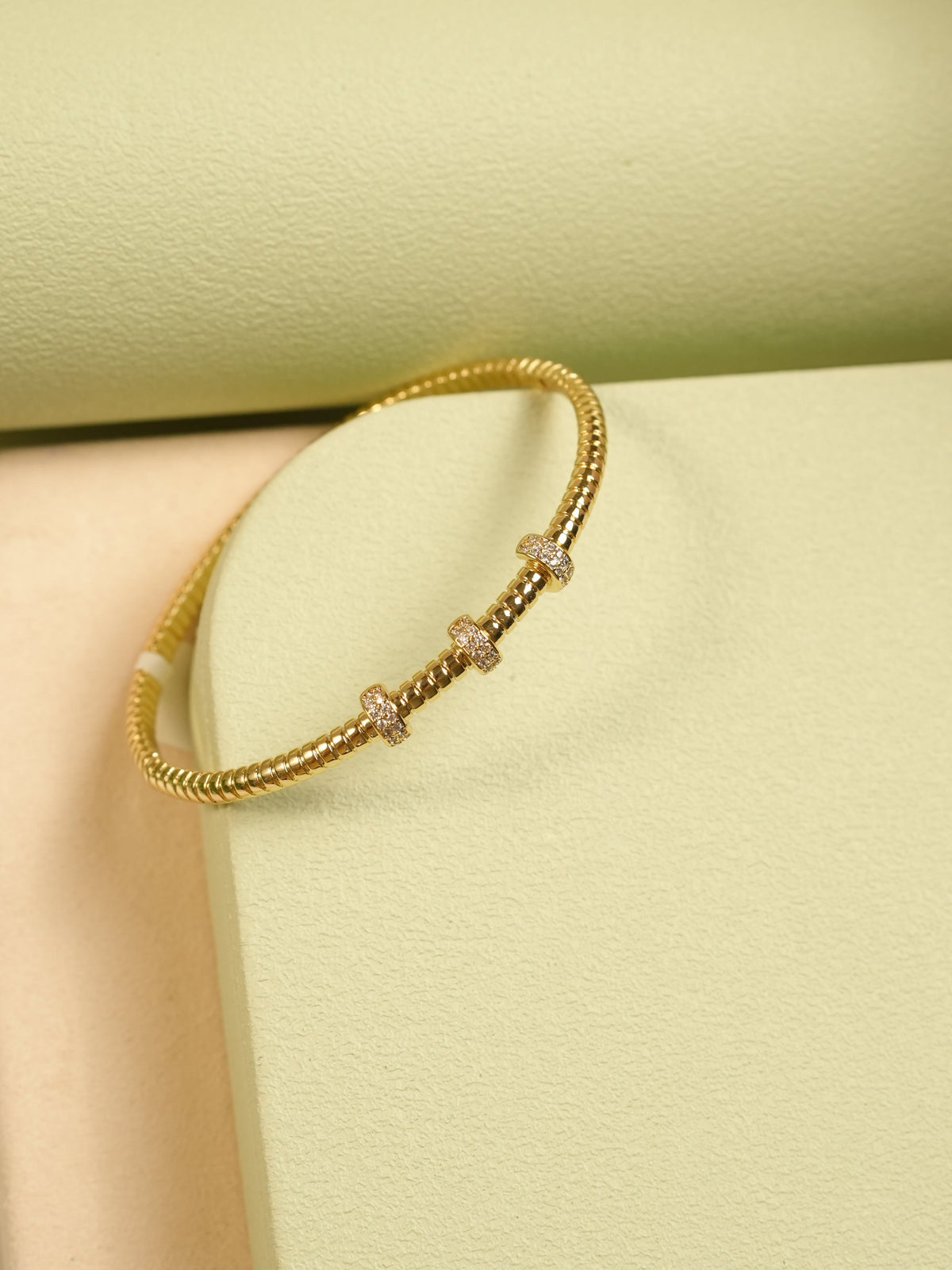 Daga-accessories gold-toned metal bangle with ribbed texture and three crystal-encrusted stations on a green display