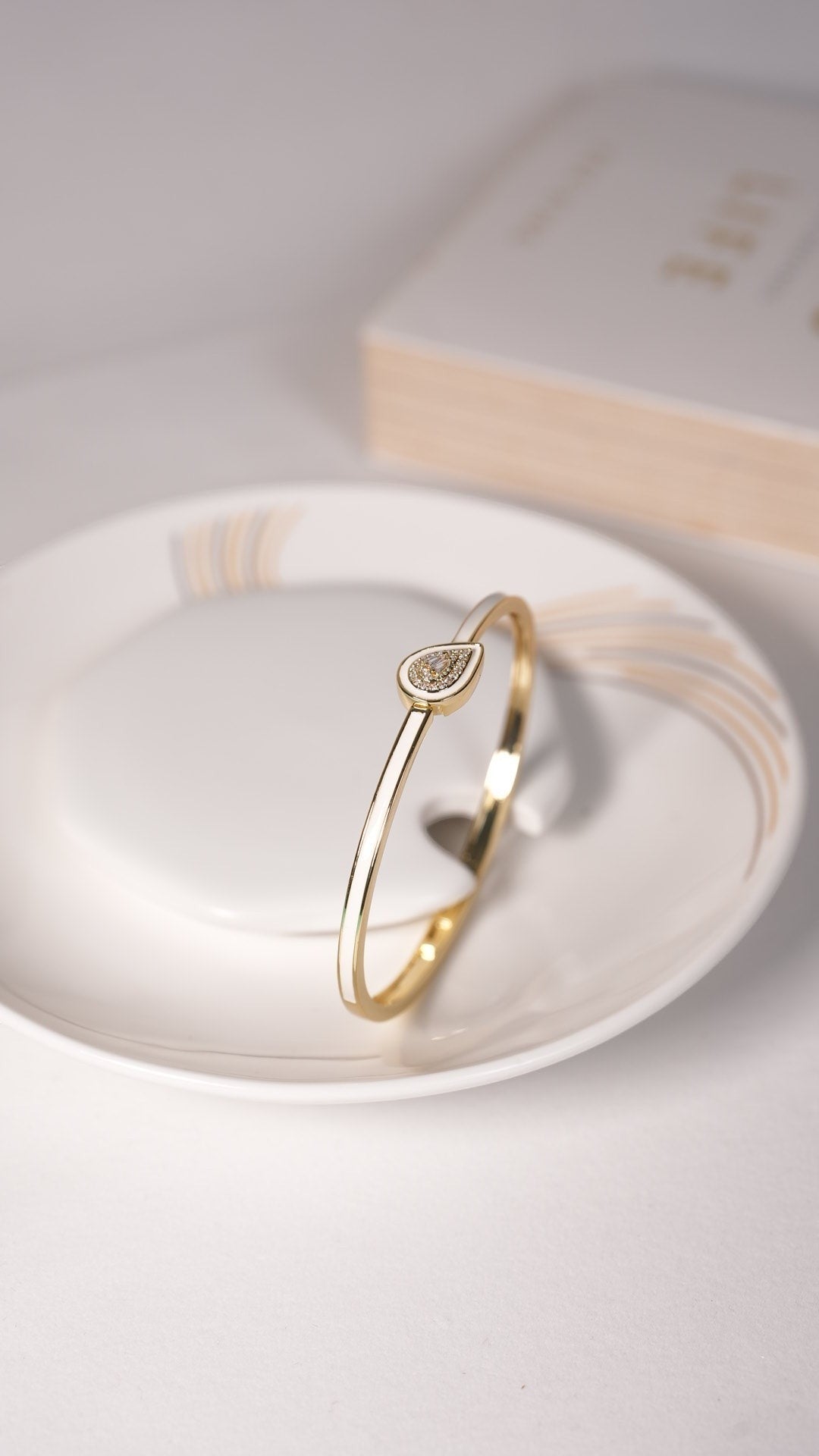 Daga-accessories ivory shine drop bracelet featuring a gold-toned band and a teardrop-shaped charm on a white dish.