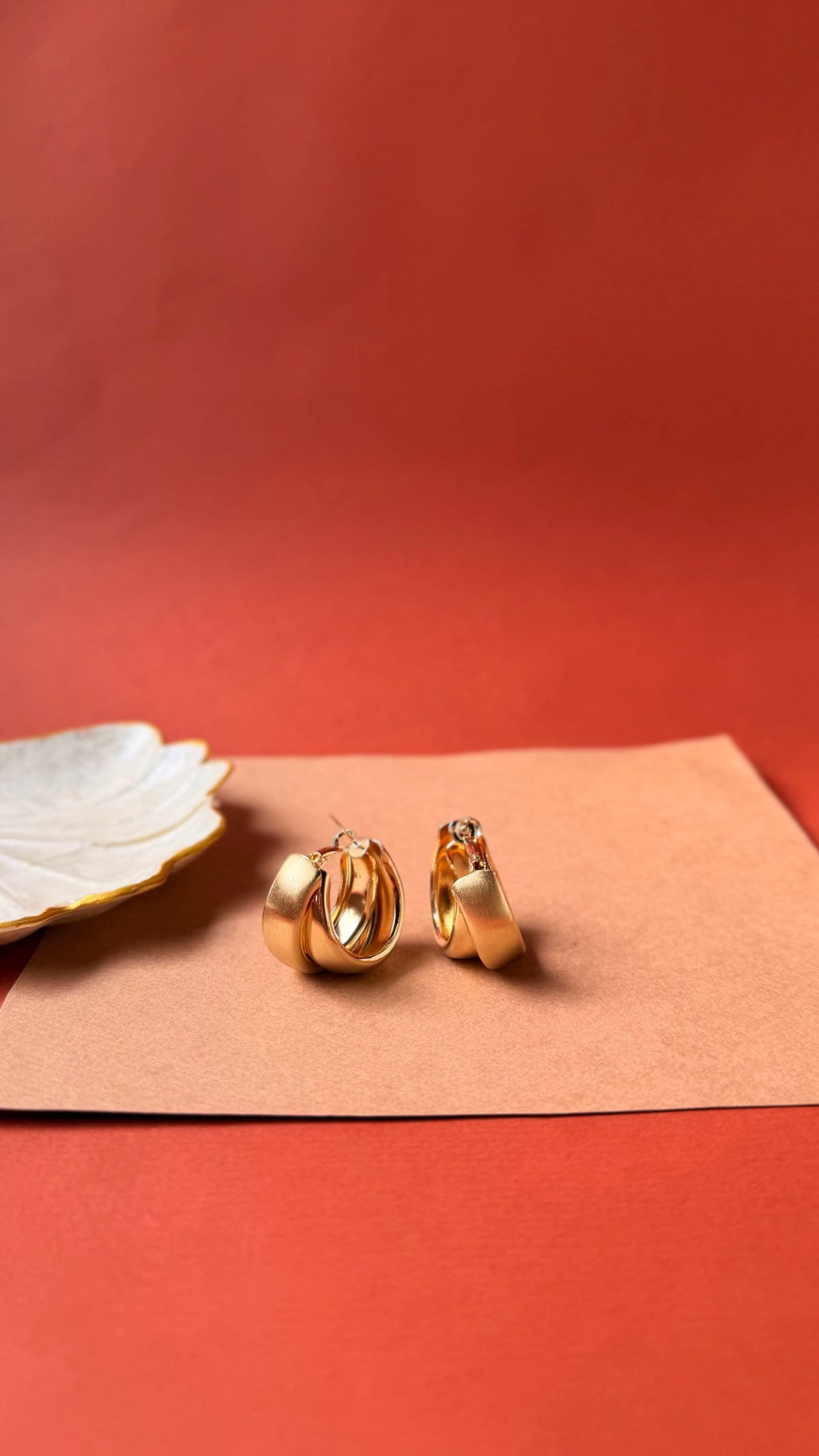 Daga-accessories Mat Orbit Gold hoop earrings with a sleek matte finish on a minimalist tan and red background.