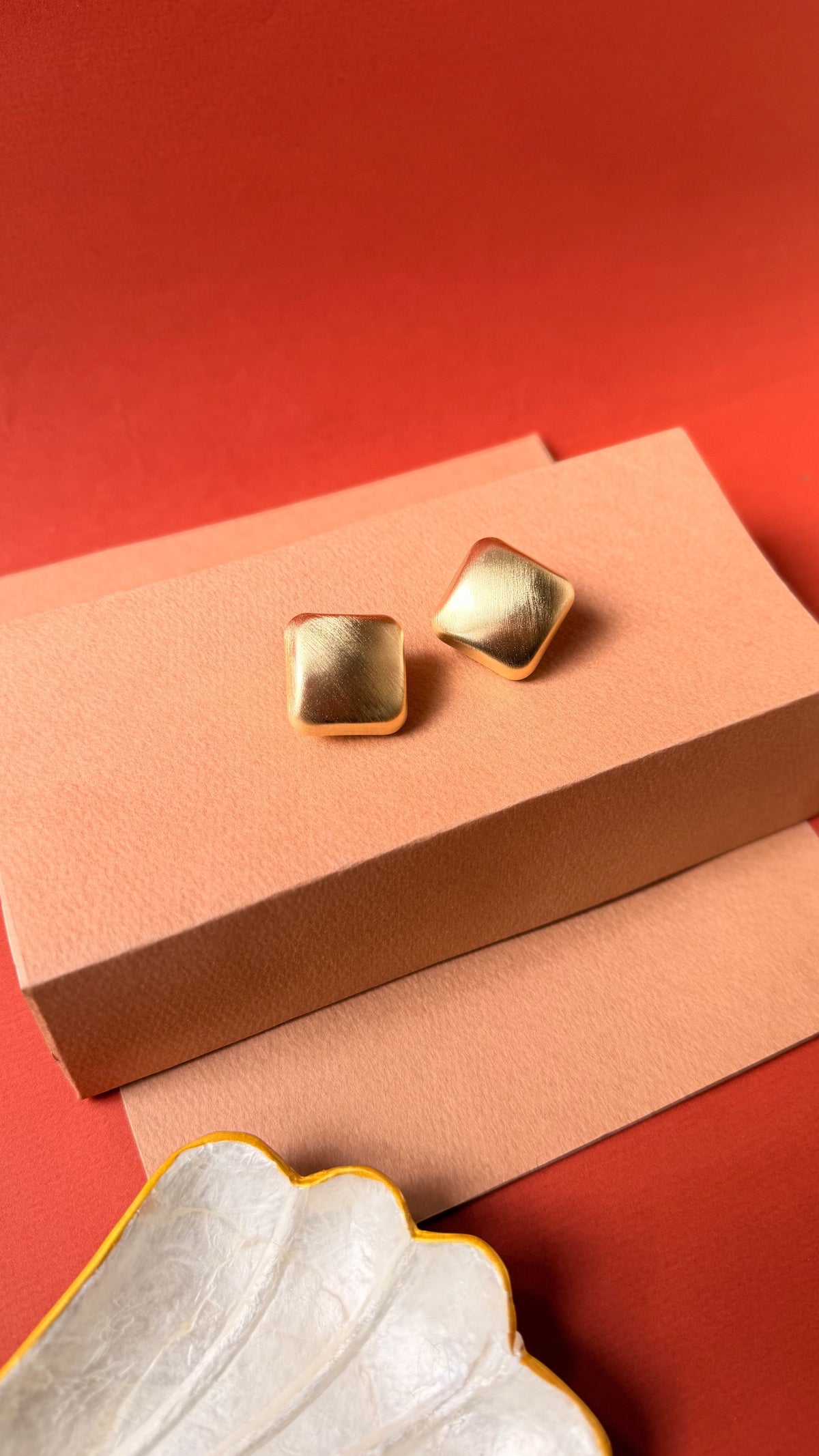 Daga-accessories matte gold square stud earrings with a modern geometric design on a peach and red background.