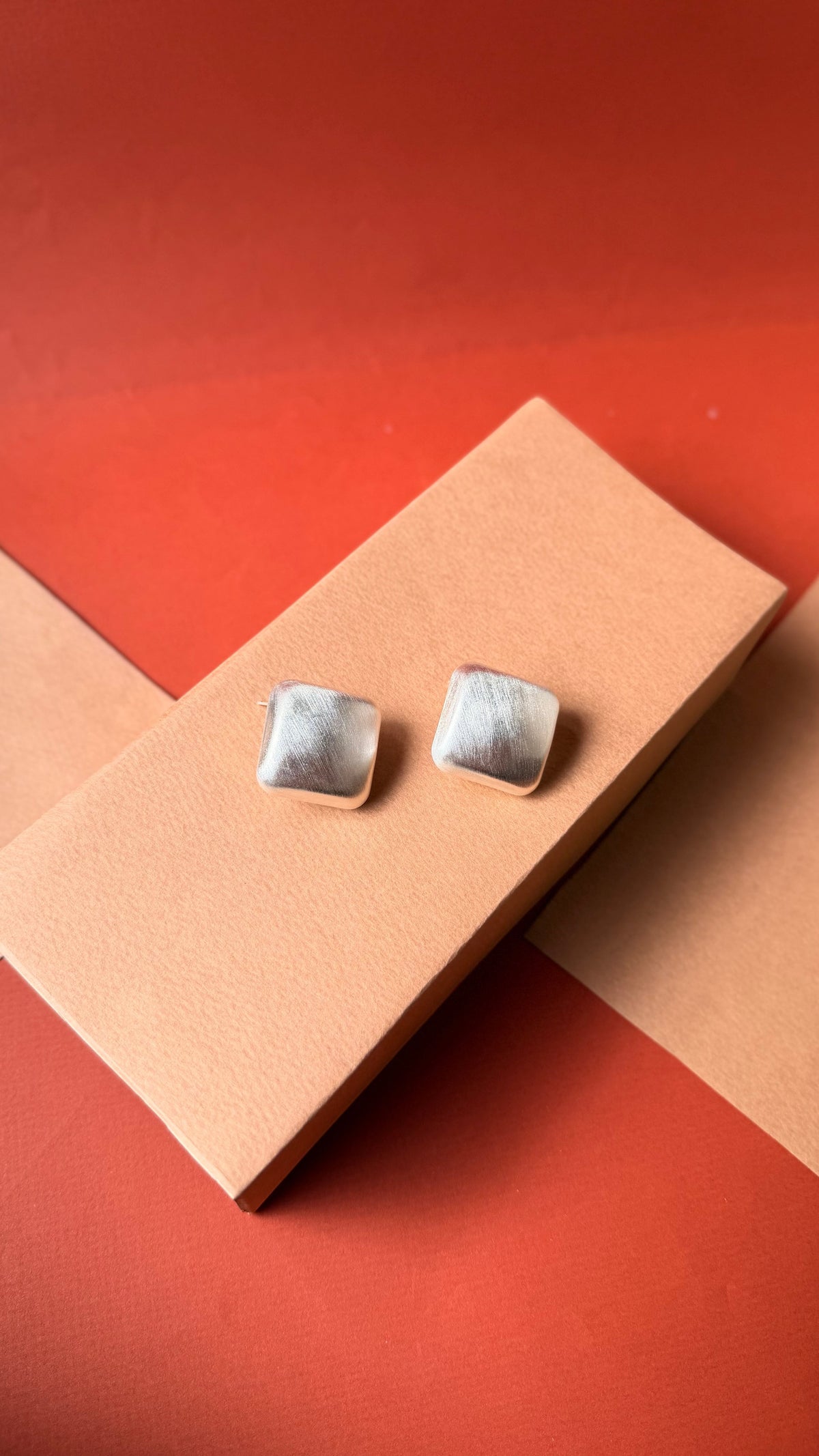 Daga-accessories matte silver square stud earrings from the Gold Bold Collection on a peach display block.