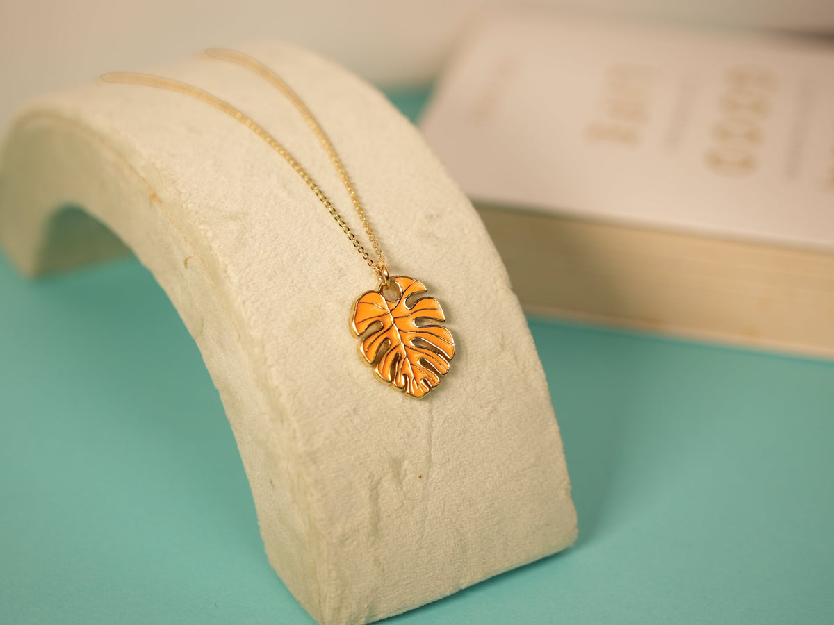 Daga-accessories gold chain necklace featuring a minimalist orange monstera leaf pendant on a stone display stand.