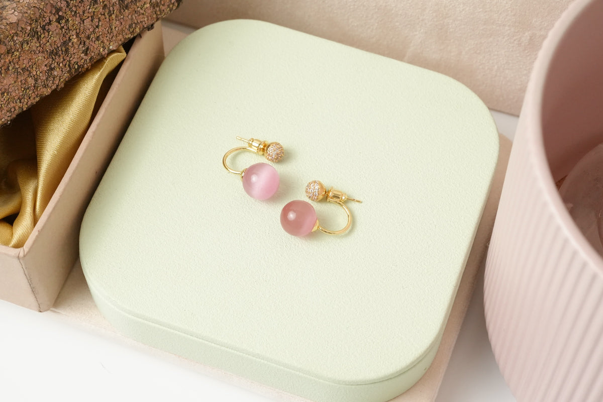 Daga-accessories gold-toned hoop earrings with pink cat-eye beads and sparkling stud details on a light green base.