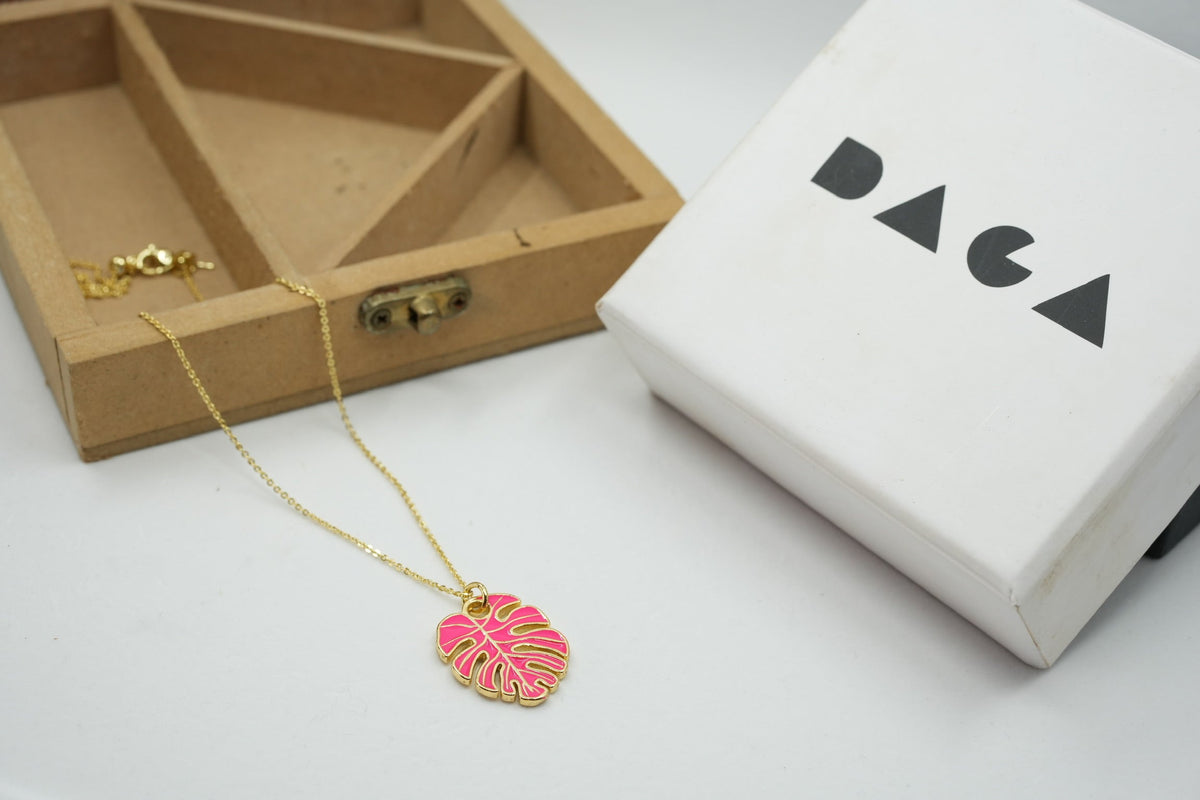 Daga-accessories Leaf of earth pink monstera leaf pendant necklace with a gold chain next to a branded gift box