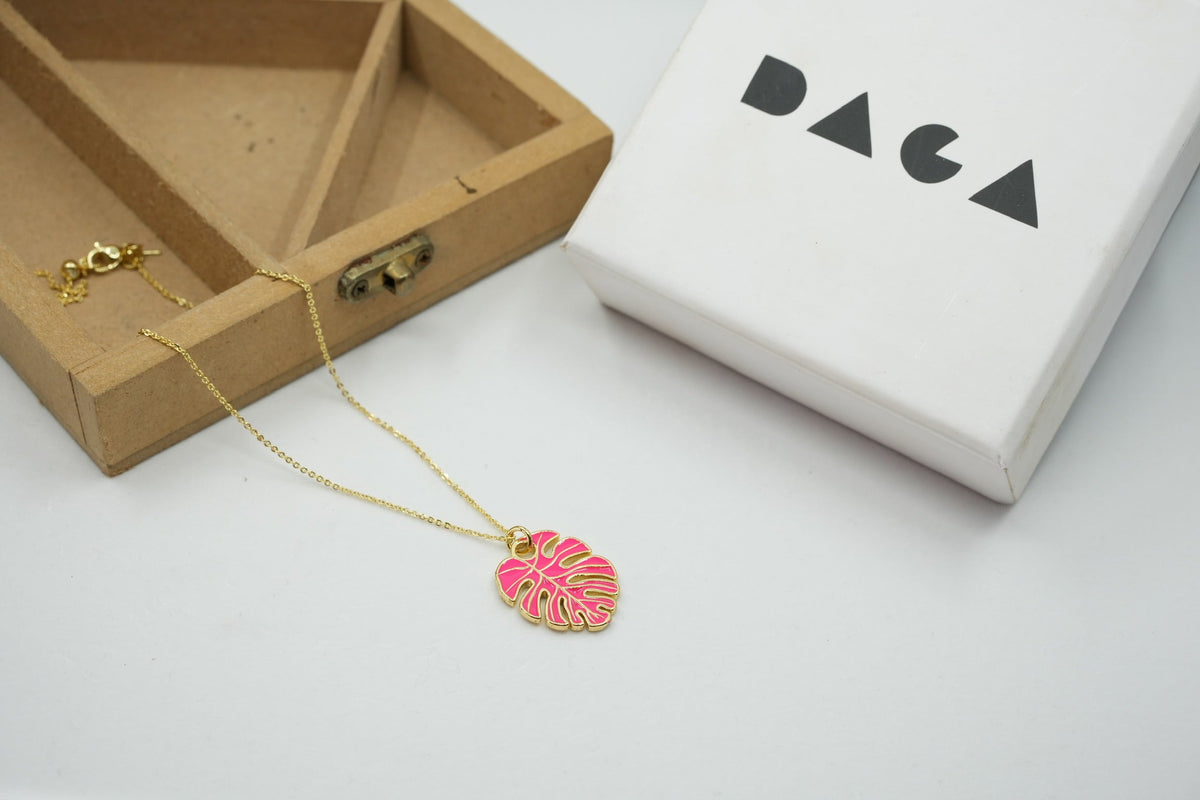 Daga-accessories gold chain necklace featuring a vibrant pink monstera leaf pendant, presented with a branded box.
