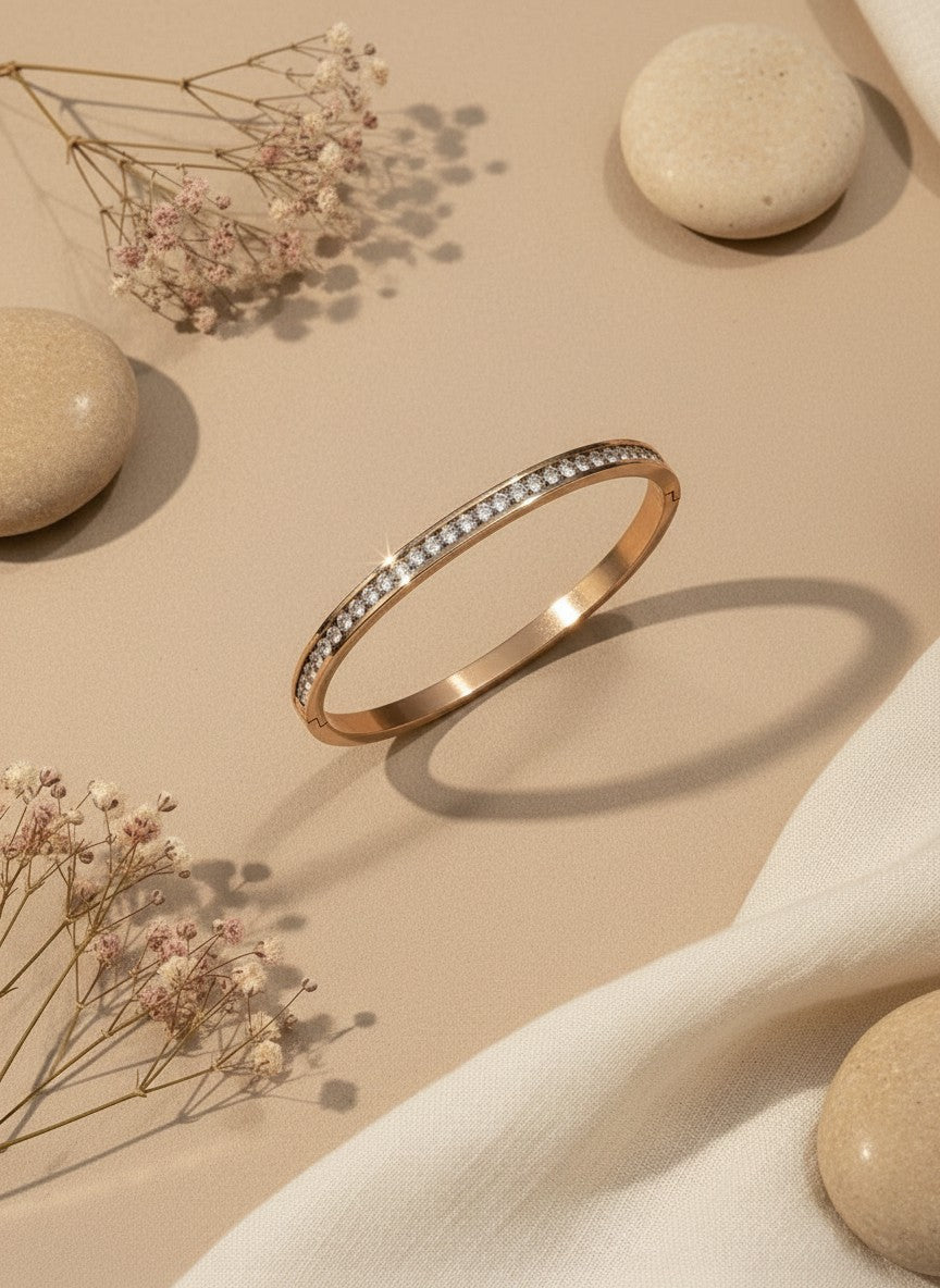 Daga-accessories rose gold-toned bangle bracelet featuring a row of shimmering crystals on a beige fabric background.