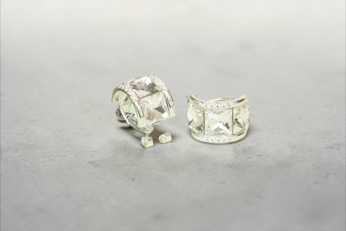 Daga-accessories silver-toned hoop earrings with square-cut crystals and halo setting on a light green display stand.