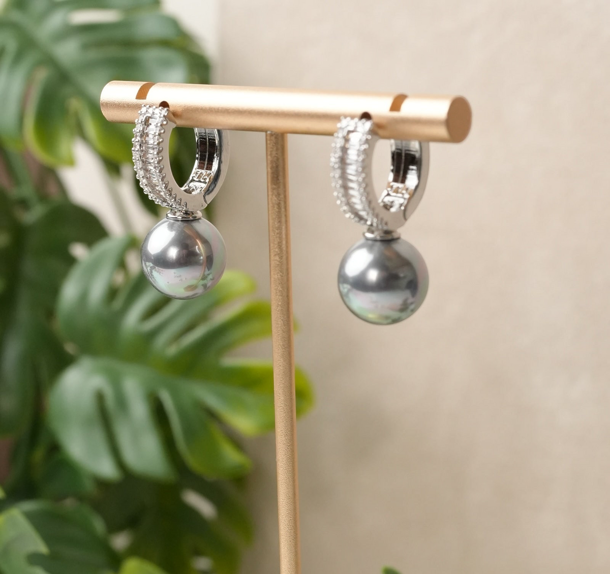 Daga-accessories silver hoop earrings with metallic-grey drop pearls and crystal-encrusted bands on a gold stand.