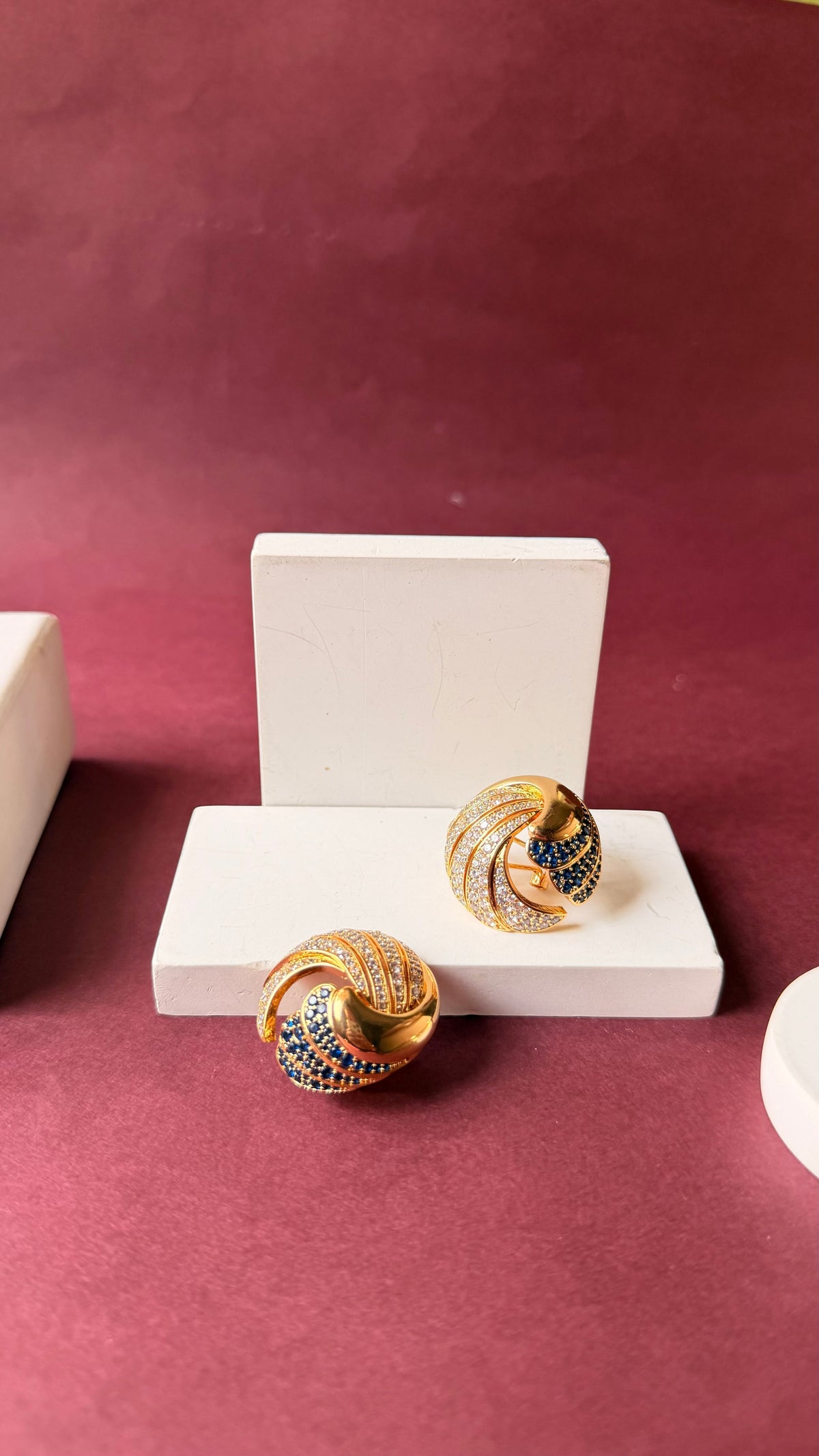 Daga-accessories Quiet spiral clip-on earrings in gold with shimmering blue and white stones on a white display stand.