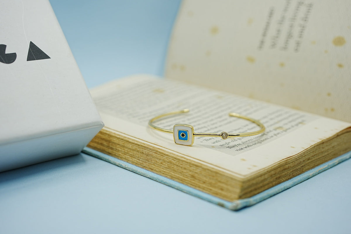 Daga-accessories gold-tone stainless steel cuff bracelet featuring a square enamel evil eye charm on an open book.