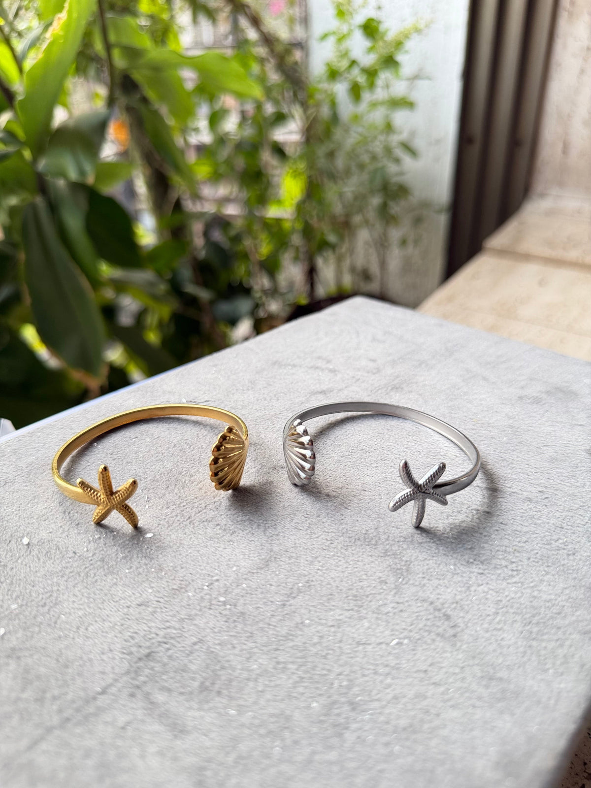 Daga-accessories gold and silver cuff bracelets featuring starfish and seashell charms on a grey display stand.