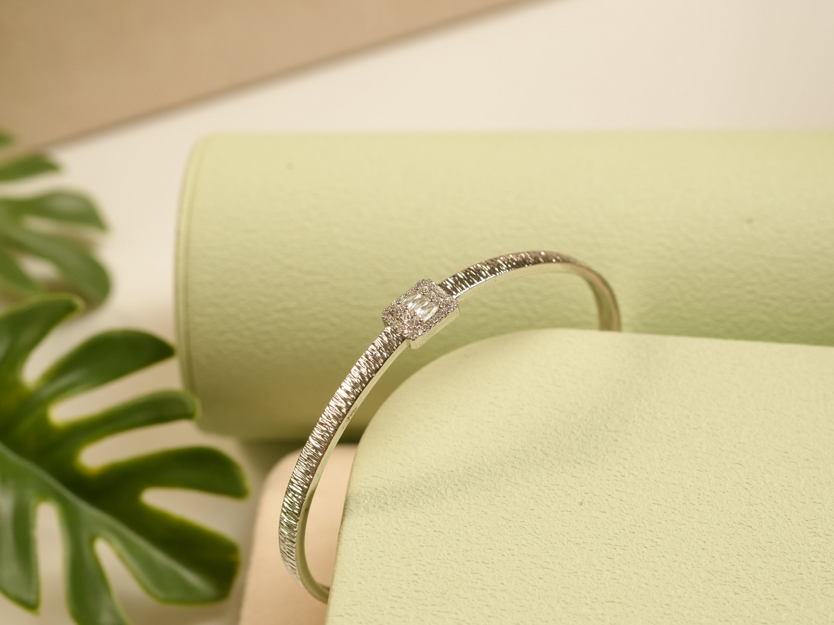 Daga-accessories textured metal bracelet with a central square stone and micro-stone frame on a green display stand.