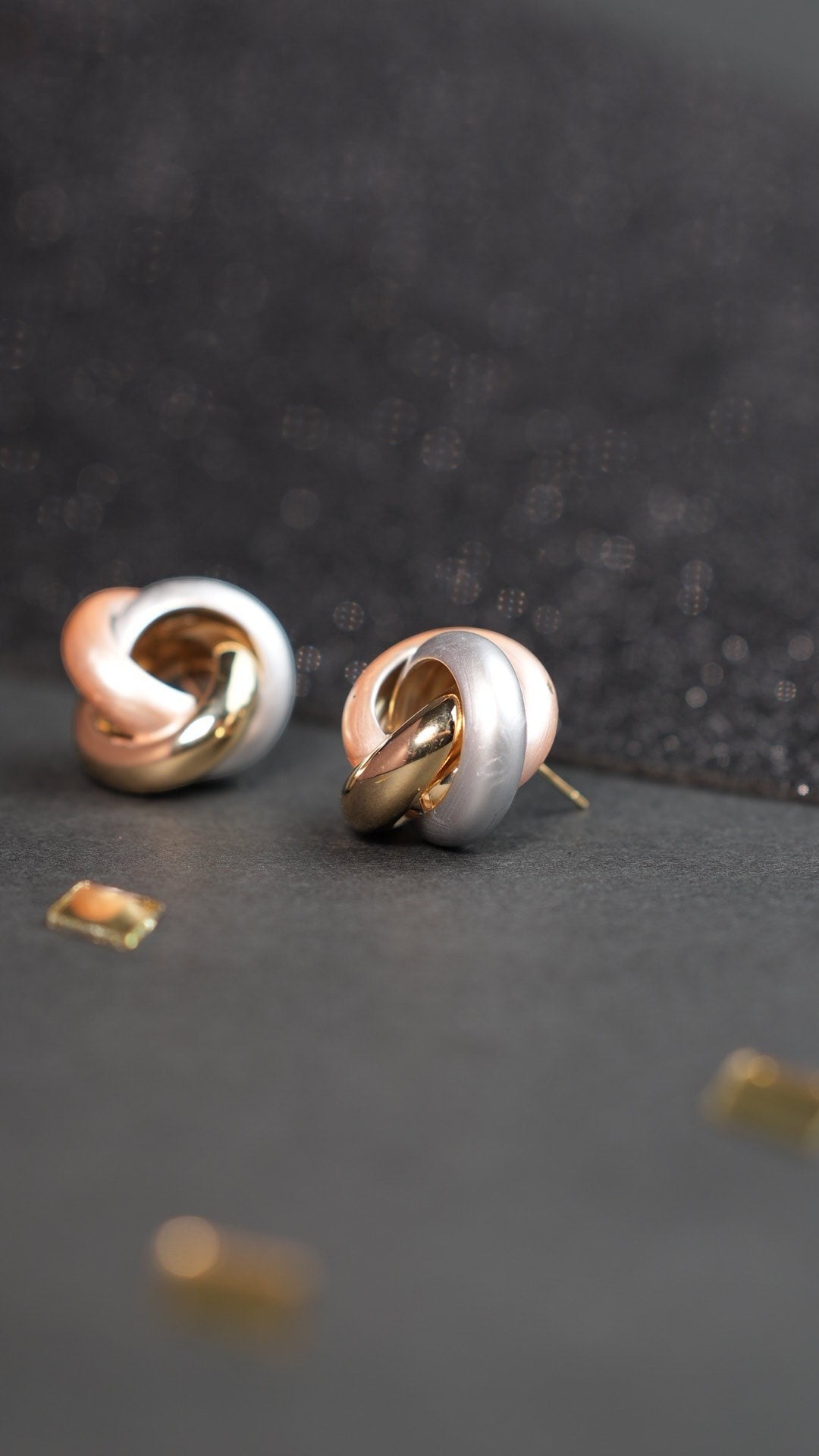 Daga-accessories 3-tone criss cross stud earrings in gold, silver, and rose gold metal on a dark textured surface.