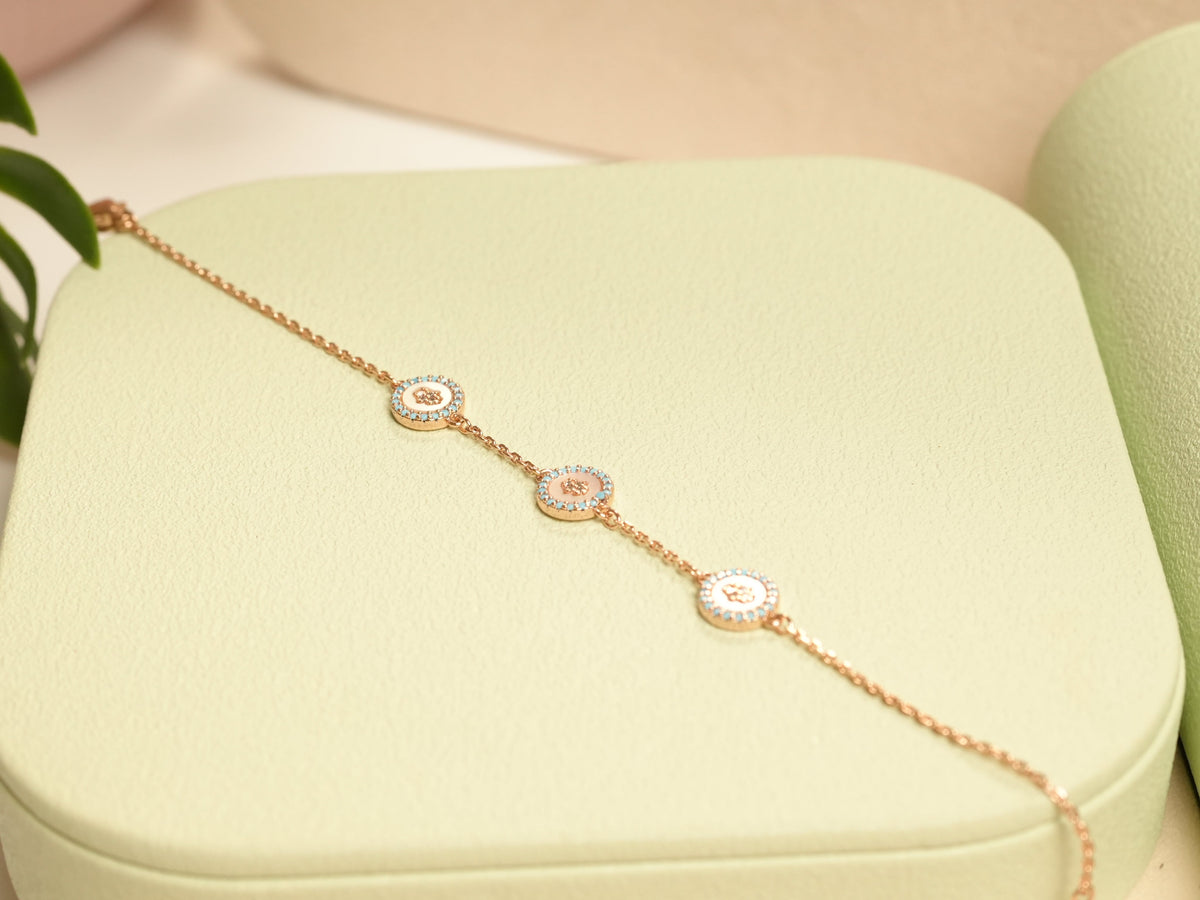 Daga-accessories rose gold triple hamsa bracelet with white enamel and blue stone accents on a light green display