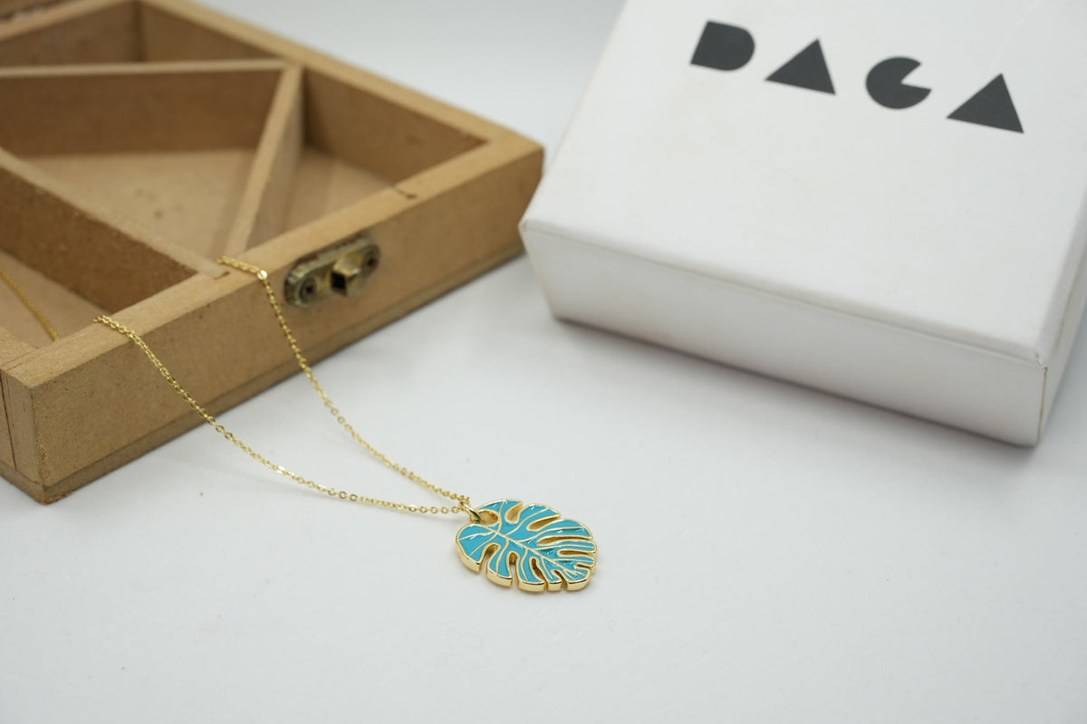Daga-accessories gold chain necklace featuring a turquoise monstera leaf pendant next to a branded gift box