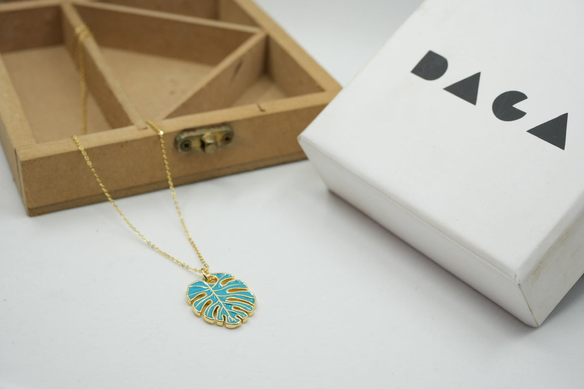 Daga-accessories gold chain necklace with a turquoise monstera leaf pendant next to a branded gift box