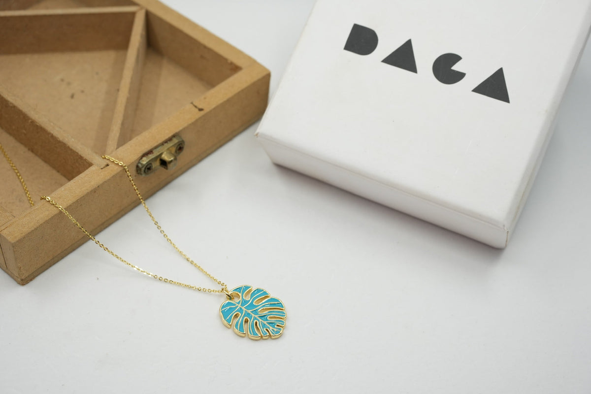 Daga-accessories gold chain necklace featuring a blue monstera leaf pendant next to a branded white gift box