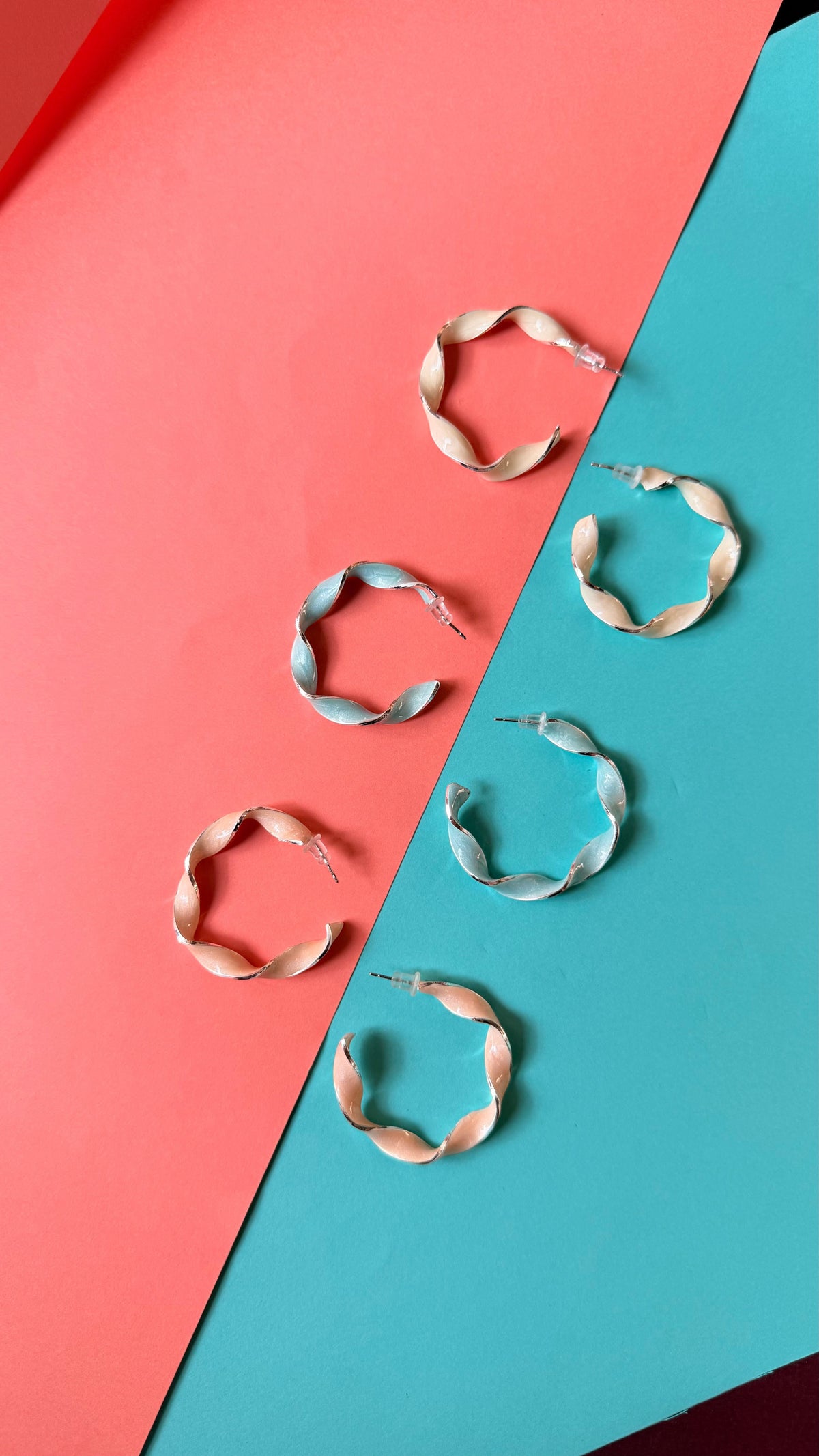 Daga-accessories twisted hoop earrings in pastel shades of peach and light blue on a split pink and teal background.