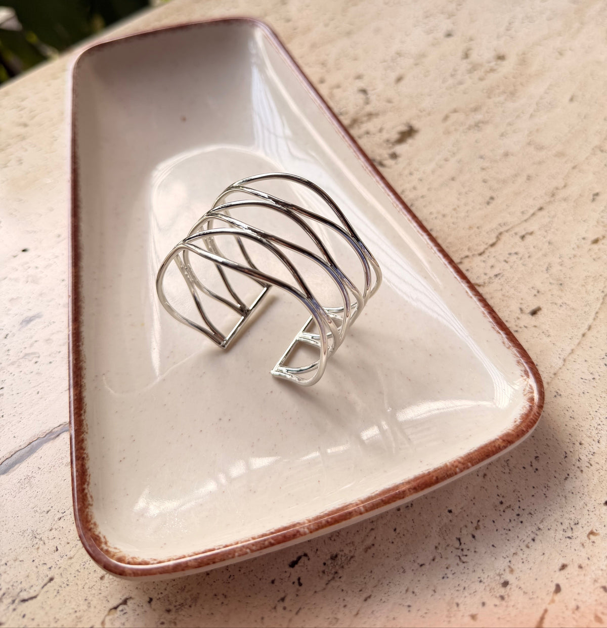 Daga-accessories wavy silver cuff bracelet with a fluid, sculptural open design resting on a ceramic tray.