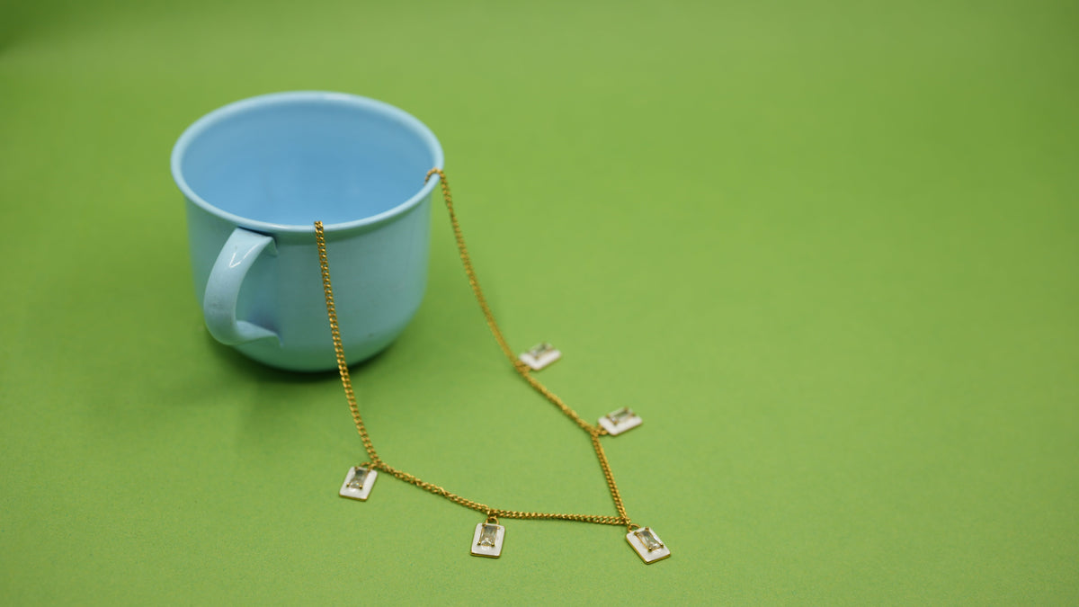 Daga-accessories white enamel rectangular charm necklace on a gold stainless steel chain draped over a blue cup.