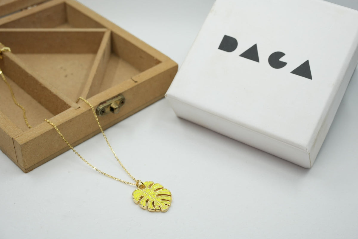 Daga-accessories yellow monstera leaf pendant necklace on a delicate gold chain next to a branded gift box
